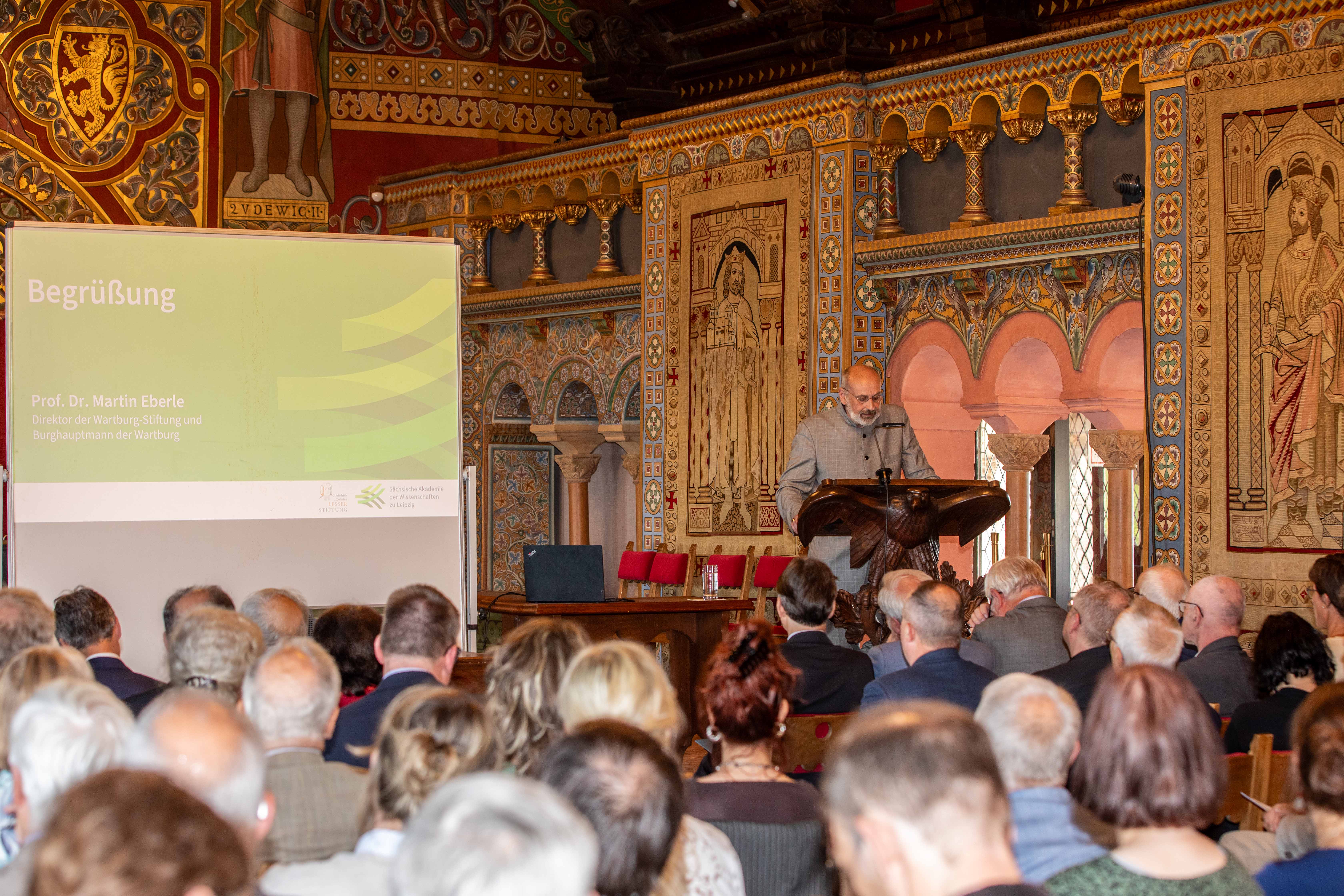 Eine männliche Person mit Brille steht in einem historischen Festsaal an einem Rednerpult und spricht in ein Mikrofon. Im Bildvordergrund sitzen Menschen auf Stühlen. Auf einer Leinwand ist zu lesen: Begrüßung Prof. Dr. Martin Eberle.