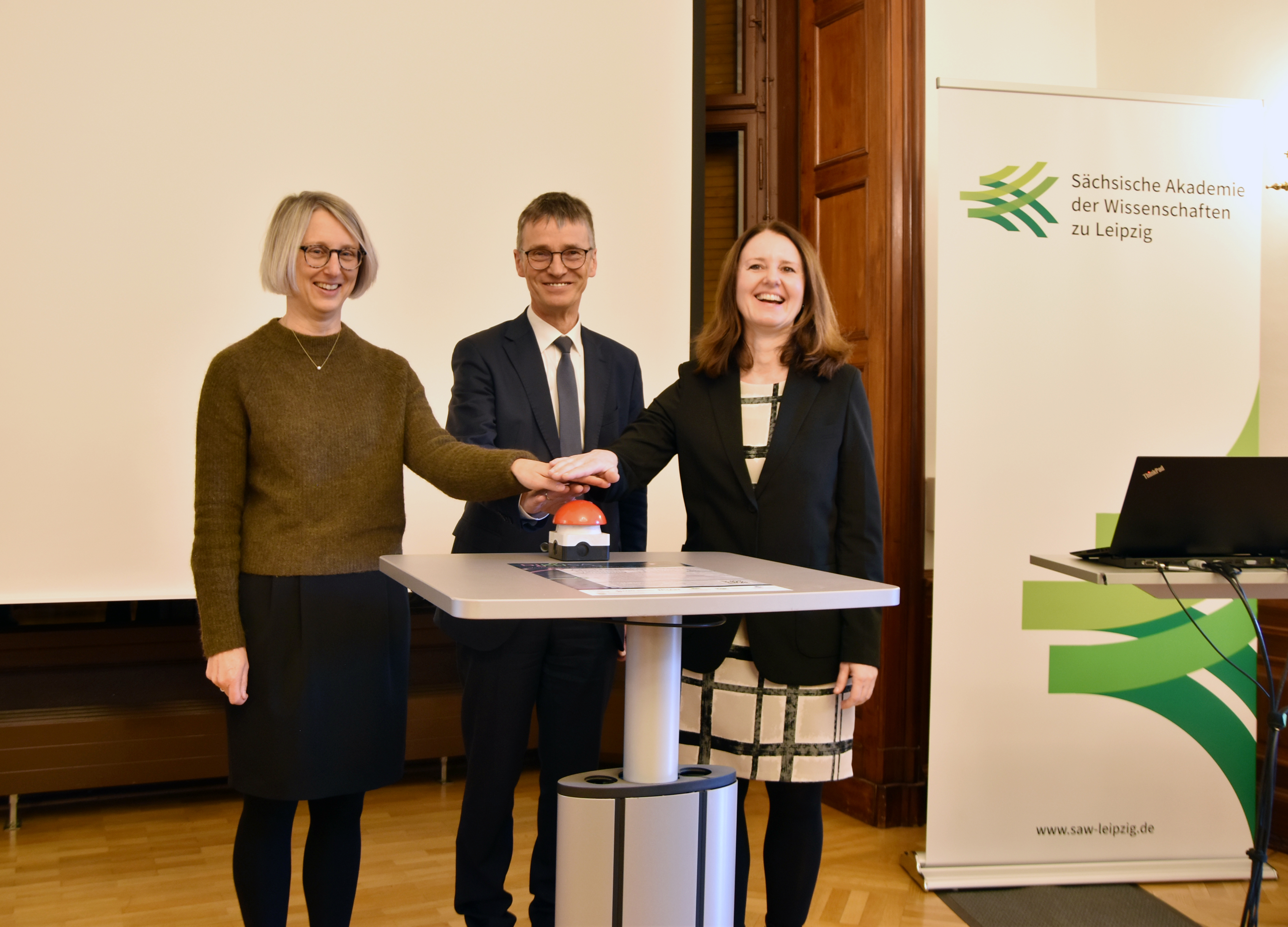 Heike Graßmann (rechts im Bild), Hans-Joachim Knölker (Mitte) und Friederike May drücken gemeinsam auf einen großen roten Knopf auf einem Tisch im Saal der Akademie. Hinter ihnen ist eine weiße Leinwand, rechts steht ein Aufsteller mit dem Logo der SAW.