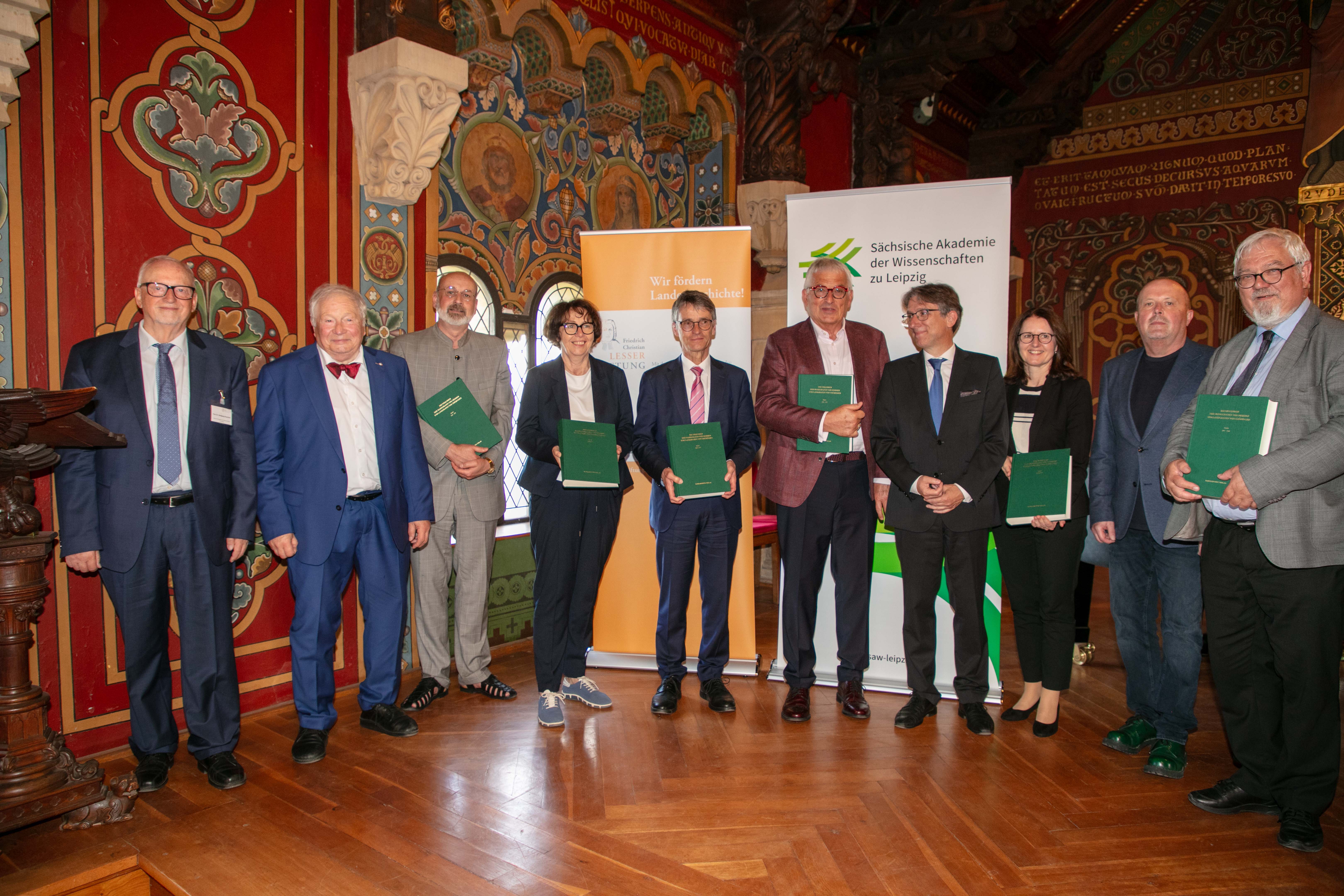 Mehrere Personen mit grünen Büchern in den Händen stehen in einem Festsaal im Halbkreis und lächeln. Im Hintergrund sieht man zwei Aufsteller; einen von der Sächsischen Akademie der Wissenschaften zu Leipzig und der Friedrich-Christian-Lesser-Stiftung