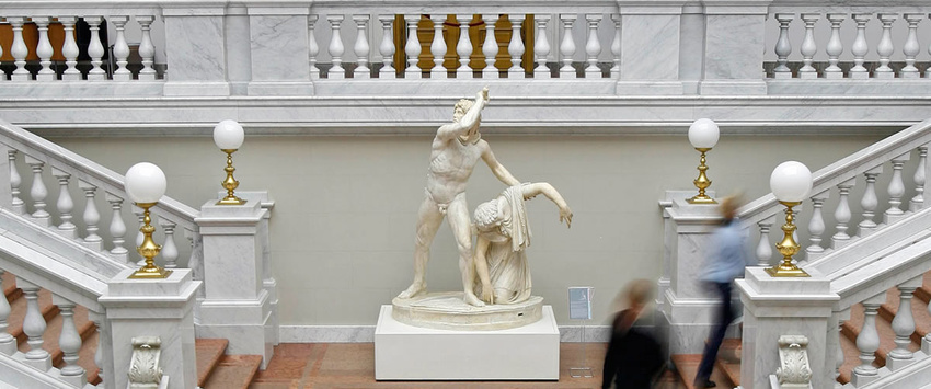 Treppenaufgänge rechts und links mit weißem steinernen Geländer und goldenen Lampen. In der Mitte des Aufgangs steht eine antike Marmor-Statue, auf der eine männliche und eine weibliche Figur zu sehen sind.