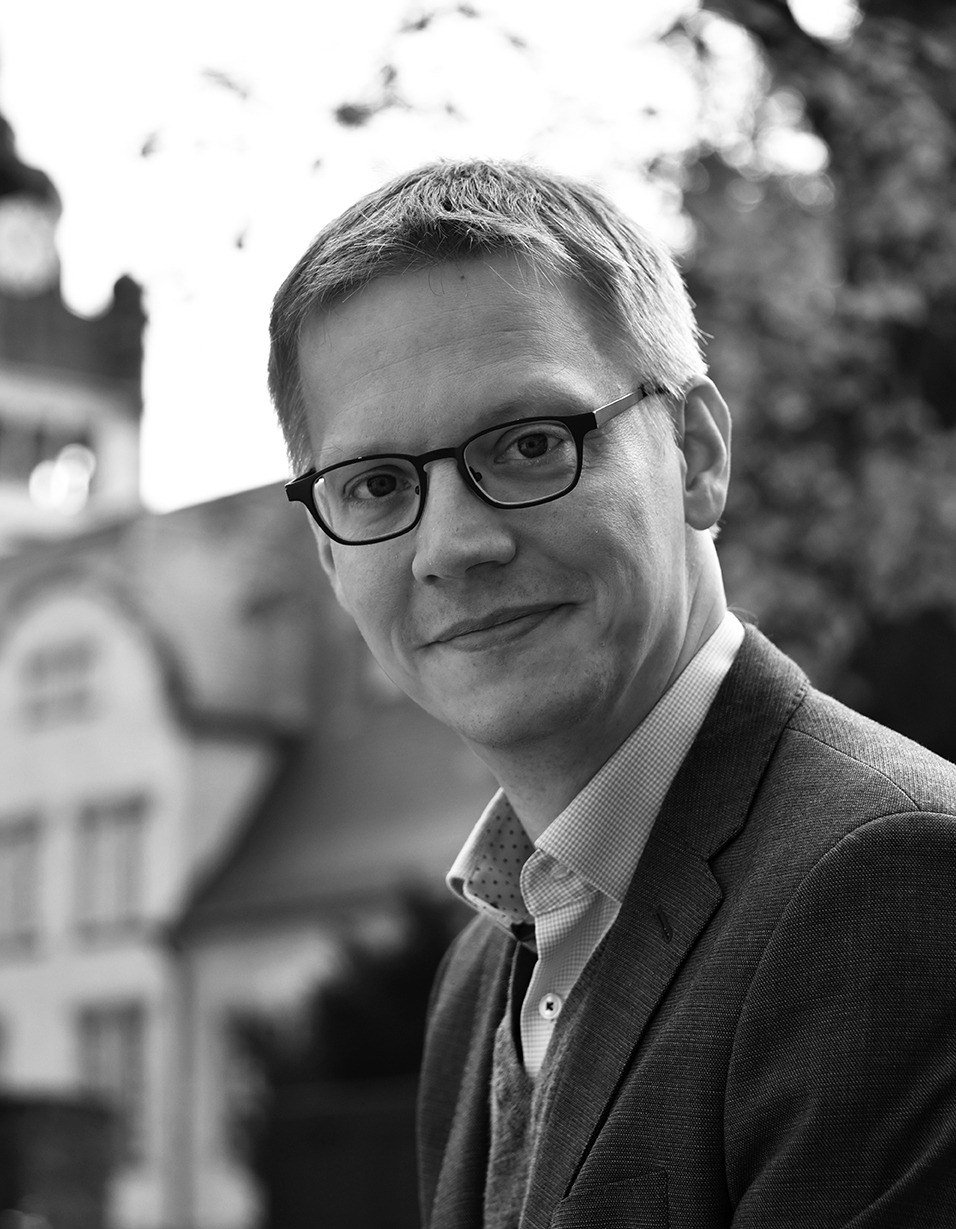Schwarz-weiß-Porträtfoto einer lächelnden männlichen Person mt Brille und Anzug vor einem historischen Gebäude
