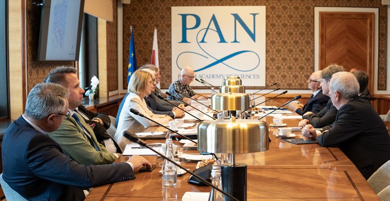 People sitting at a conference table with lamps and drinks in the middle of the table. On the wall at the back there is a poster with PAN in big letters