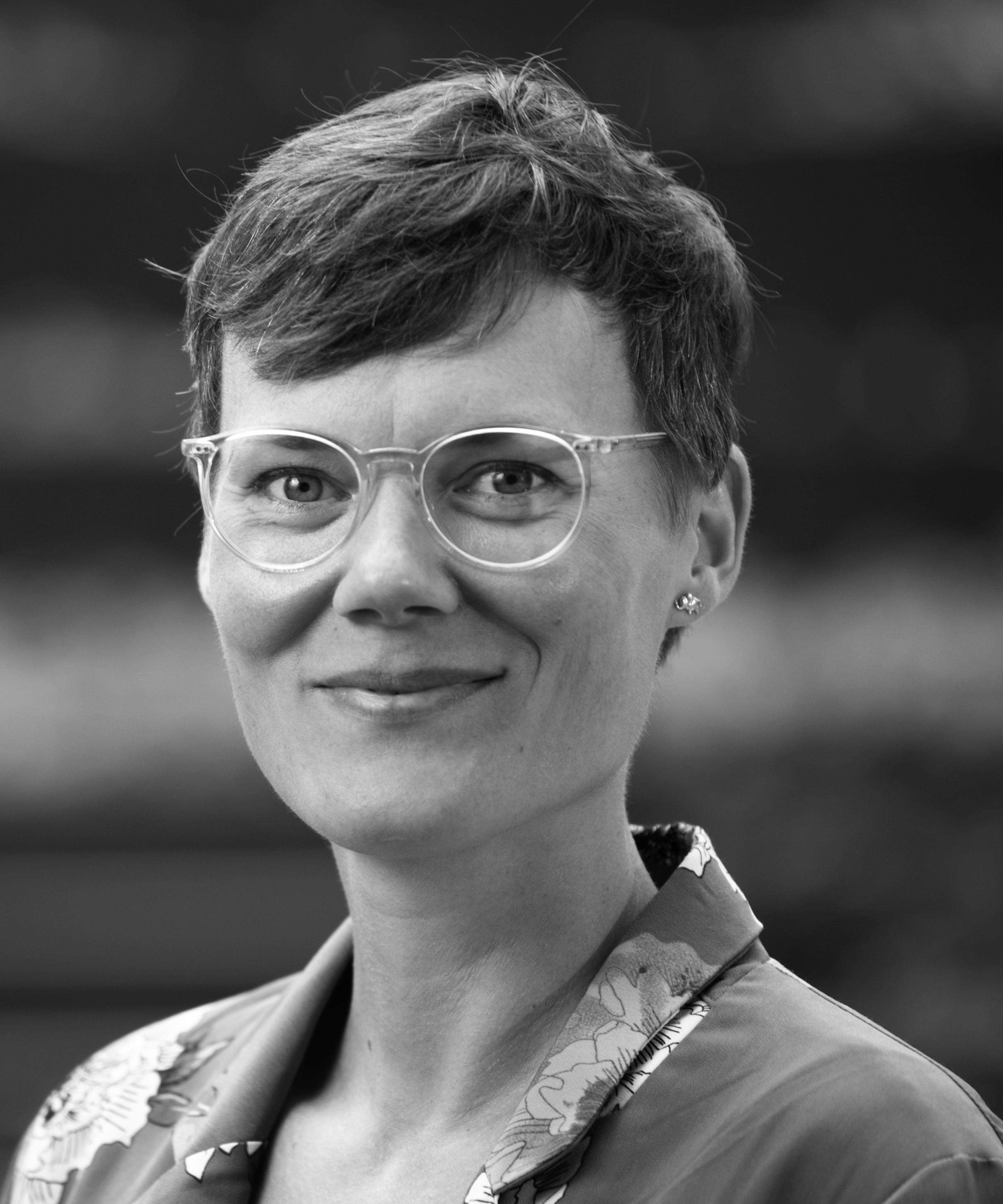 A black-and-white portrait photograph of a smiling woman wearing glasses; at the bottom of the image is a green graphic element bearing the inscription ‘Junges Forum’